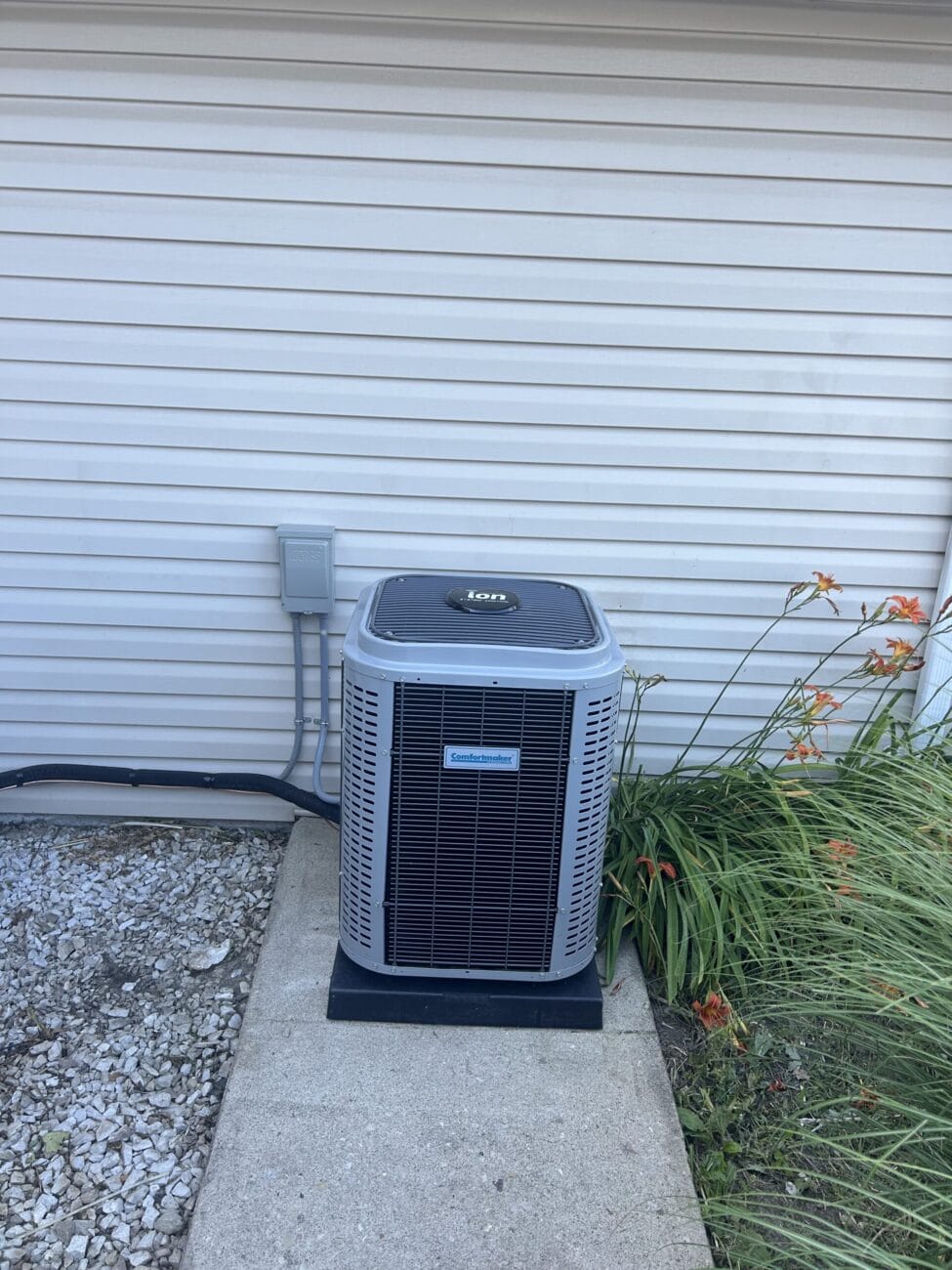 An outdoor air conditioning unit sits on a concrete pad next to a house with vinyl siding and some tall orange flowers nearby.