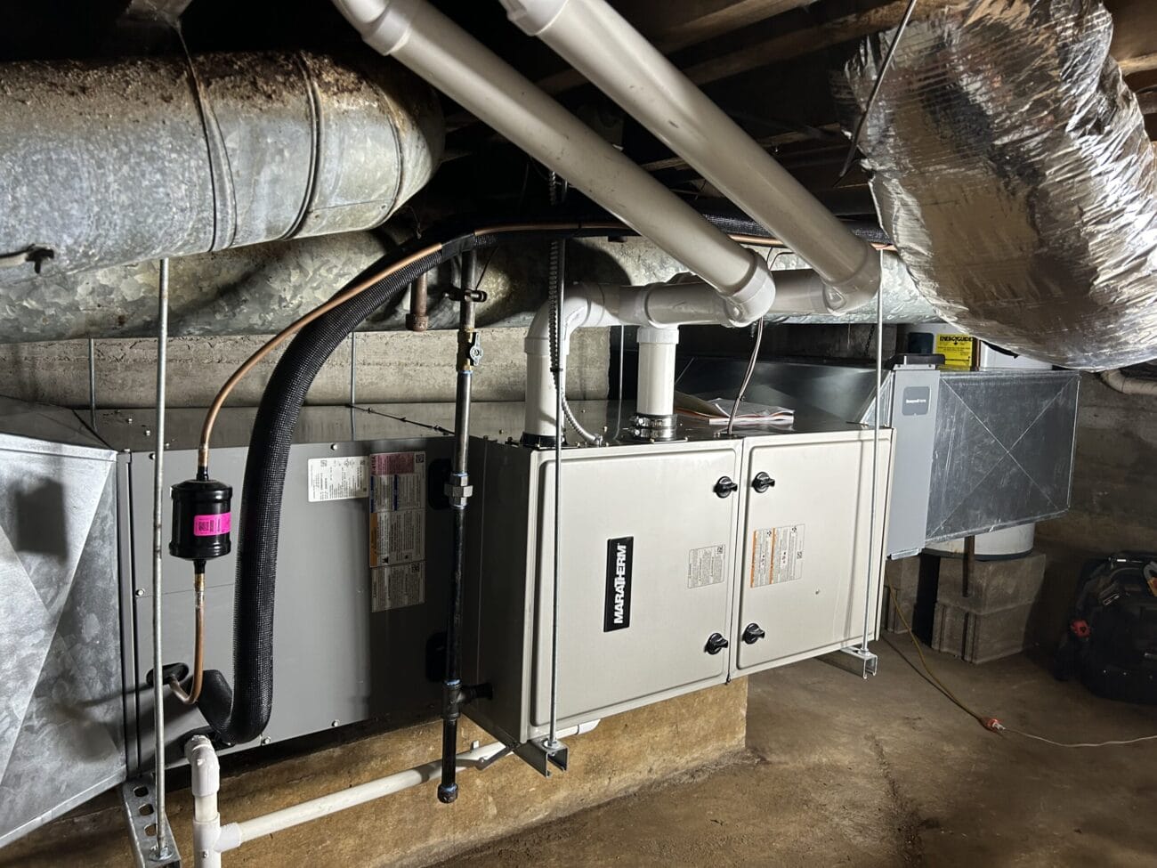 A residential basement HVAC system with ducts, pipes, and insulation is installed along the ceiling and walls. The area has concrete floors and unfinished walls.
