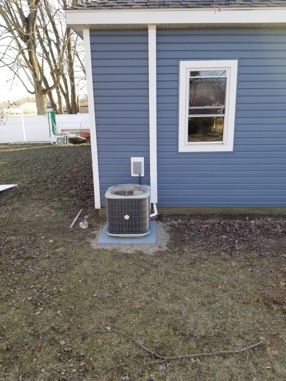 An outdoor air conditioning unit sits on a concrete pad next to a blue house with white trim and a single window.
