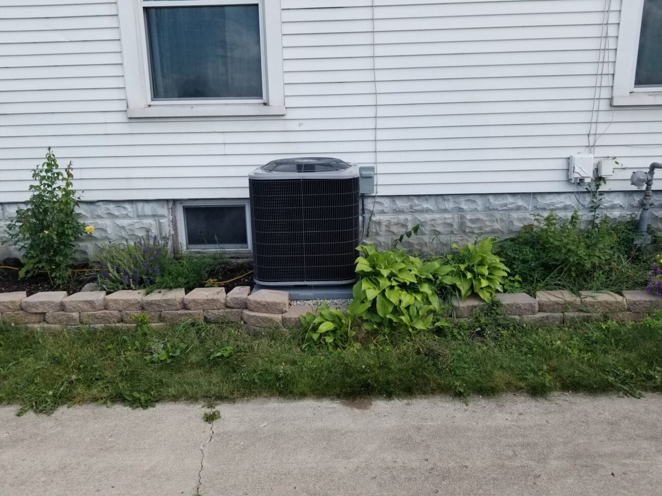 An outdoor air conditioning unit is installed next to a white house, surrounded by a small garden with plants and a stone border.