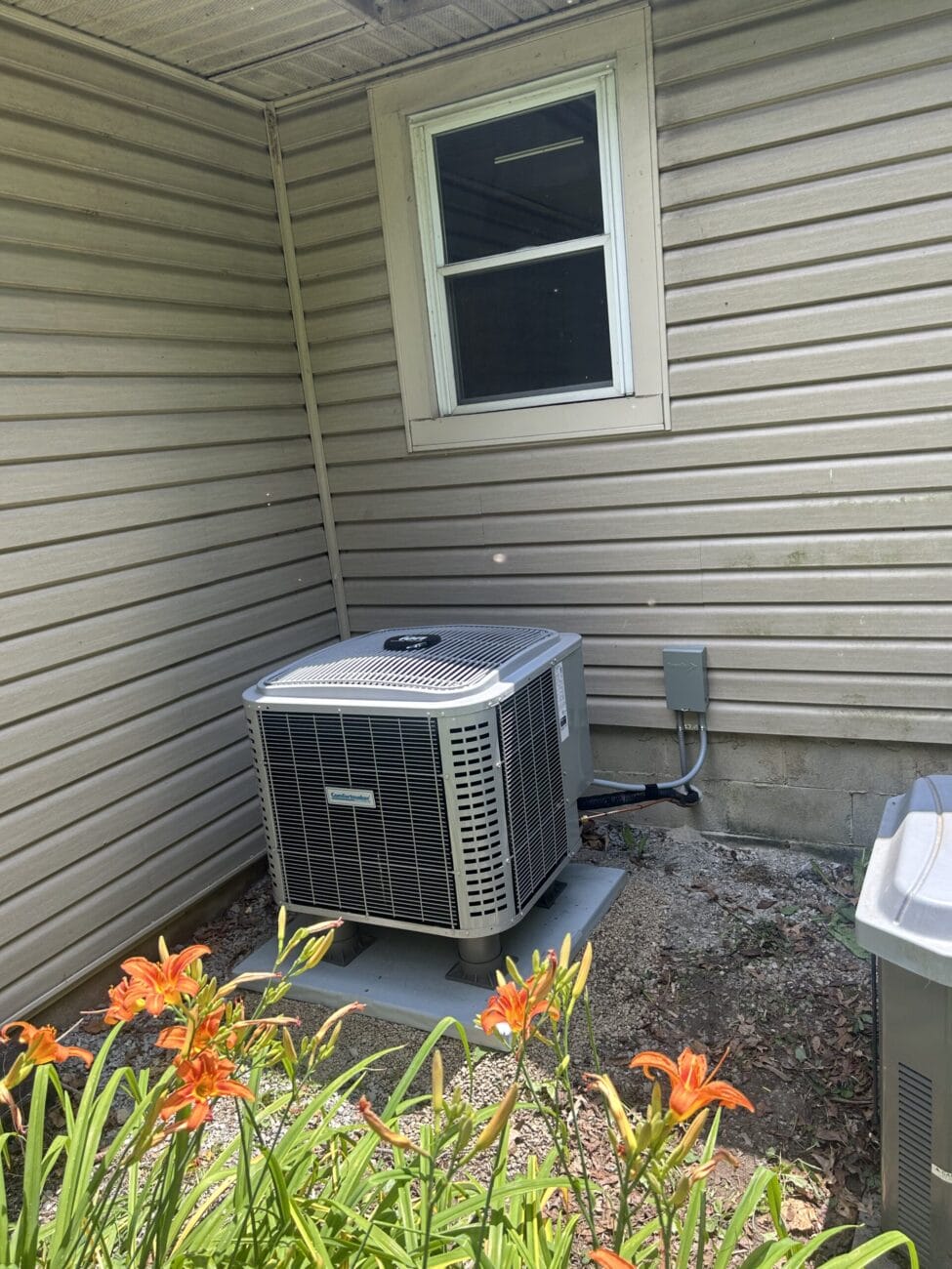 Outdoor air conditioning unit installed next to a house with beige siding, a small window above, and orange lilies in the foreground.