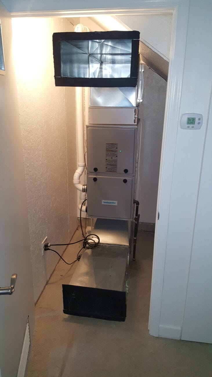 A residential furnace unit installed in a small utility closet with visible ductwork, thermostat on the wall, and electrical cords on the floor.
