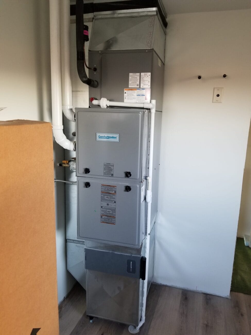 A gray central heating furnace unit installed in a residential utility room with pipes and ducts connected.