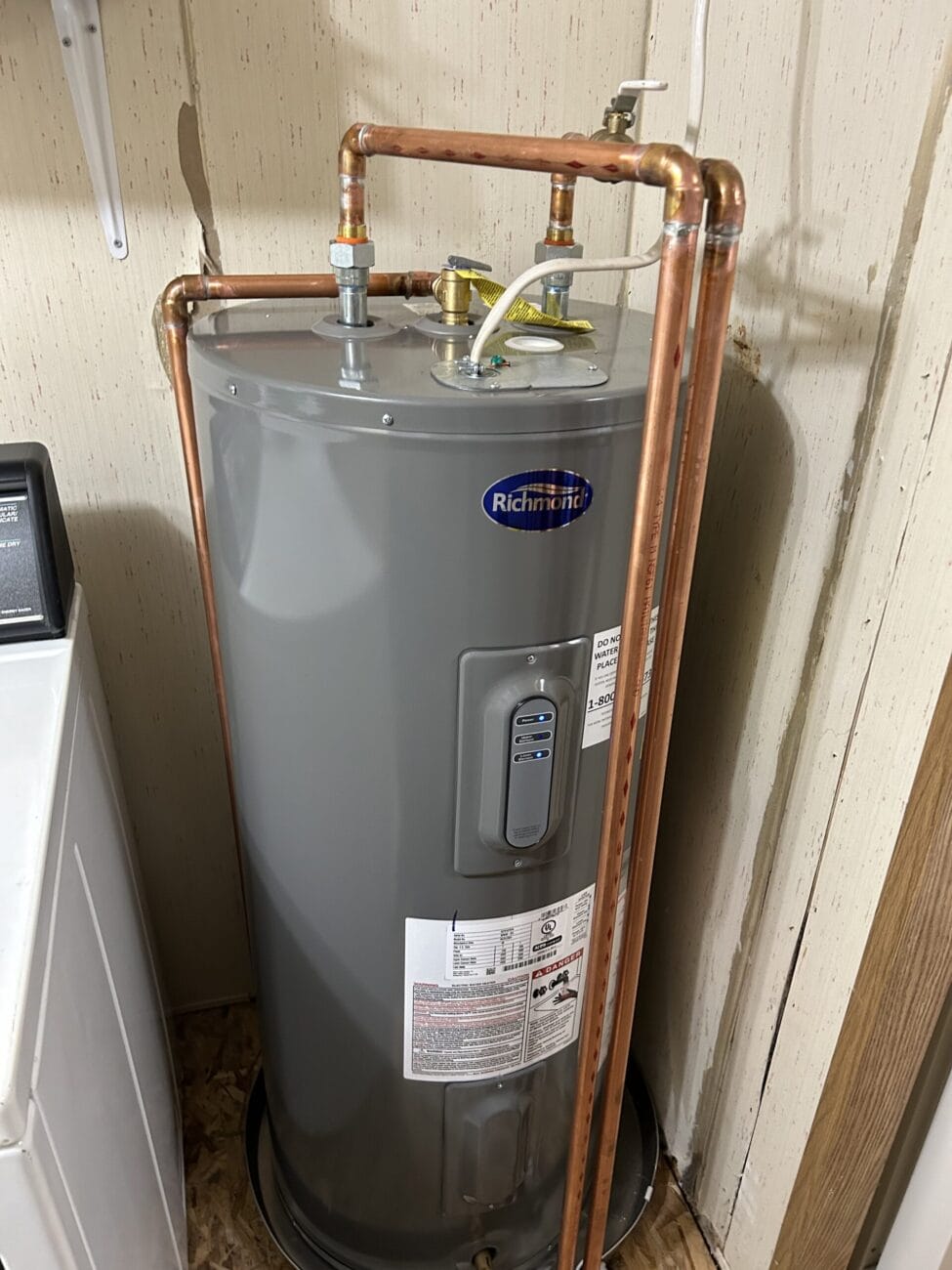 A Richmond water heater with copper piping installed in a corner next to a washing machine in a utility room.