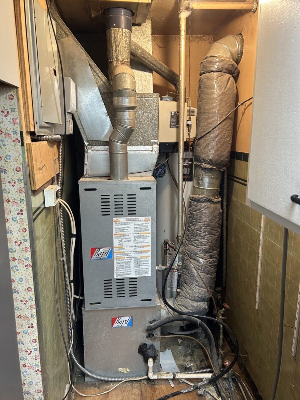 A central heating furnace with ductwork and insulated pipes is installed in a narrow utility closet with green tile walls and visible wiring.